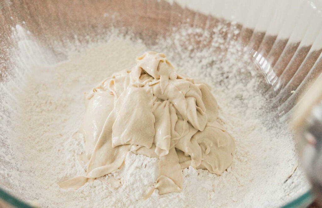 A large glass bowl contains a mound of creamy hi-protein yogurt placed on a bed of white flour. The mixture appears smooth and ready for kneading or further preparation. The transparent bowl shows a hint of a wooden surface underneath.